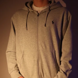 Grå ziphoodie från Ralph Lauren - Säljer en stilren grå hoodie från Ralph Lauren med dragkedja och justerbar huva. Perfekt för en avslappnad look. Den har långa ärmar och en liten logotyp på bröstet.