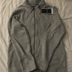 Ljusgrå kofta från Jack & Jones - Säljer en stilren ljusgrå kofta från Jack & Jones med dragkedja och klassisk krage. Perfekt för lager-på-lager under kyligare dagar. Koftan är tillverkad i ett mjukt material som ger en bekväm passform.