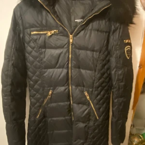 Svart quiltad jacka med pälsdetaljer - Snygg svart quiltad jacka med dragkedja och pälsdetaljer på huvan. Jackan har flera praktiska fickor med dragkedjor och en elegant design. Perfekt för kyligare dagar.