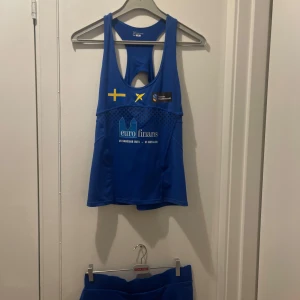 Blå linnetopp från Craft - Säljer en blå linnetopp  med tryck av svenska flaggan och texten 'euro finans'. Toppen har en brottarrygg och är perfekt för träning. Med en matchande shorts tjol 