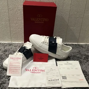 Valentino Garavani sneakers - Snygga vita Valentino Garavani sneakers med breda mörkblå remmar över ovandelen. Skorna har en klassisk design med snörning och är tillverkade i skinn. Perfekta för en stilren look.