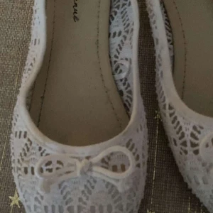 Vita spetsballerinaskor - Säljer ett par eleganta vita ballerinaskor med spetsdetaljer och en söt rosett framtill. Perfekta för en somrig look eller en speciell tillställning.