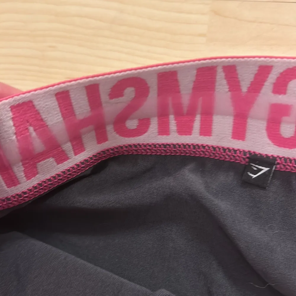 Snygga svarta shorts från Gymshark med en bred, elastisk midja i rosa och vit med logotyp. Perfekta för träning och bekväma att bära. Tyvärr är lappen bortklippt men det är storlek S. Shortsit.