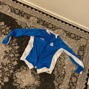 Blå och vit jacka från Espanyol de Barcelona Academy - Snygg blå och vit jacka med dragkedja från Espanyol de Barcelona Academy. Perfekt för sportiga aktiviteter med sitt lätta och bekväma material. Jackan har långa ärmar och en stilren design med logotyp på bröstet och ryggen.