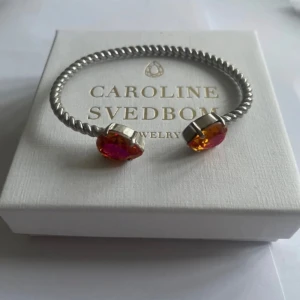 Armband med färgglada stenar från Caroline Svedbom - Elegant armband från Caroline Svedbom med en tvinnad silverfärgad design. Armbandet pryds av två stora, färgglada stenar i orange och rosa nyanser som ger en lyxig känsla. Perfekt för att lägga till en färgklick till din outfit. Box ingår ej.