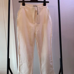 Vita byxor från NA-KD  - Stilrena vita byxor från na-kd x Angelica Blick med hög midja och rak passform. Byxorna har en elegant design med diskreta fickor och är perfekta för en chic look. Tillverkade i ett bekvämt material som ger en snygg siluett.