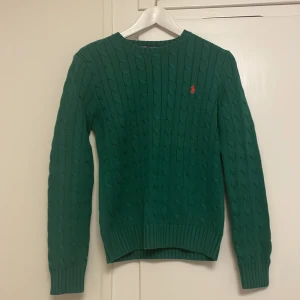 Grön stickad tröja från Ralph Lauren - Säljer en snygg grön stickad tröja från Ralph Lauren med kabelstickat mönster och det klassiska röda logotypen på bröstet. Tröjan har långa ärmar och ribbade muddar för en bekväm passform. Perfekt för kyligare dagar!