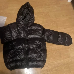 Svart pufferjacka från Axel Luxe - Snygg svart pufferjacka från Axel Luxe med huva och dragkedja. Perfekt för kyliga dagar med sin vadderade design som håller dig varm. Jackan har en glansig finish och är idealisk för både stil och komfort. Köpte den för nån vecka sen och den passade inte mig tyvärr så jag säljer den lite minde än vad ja köpte den för 