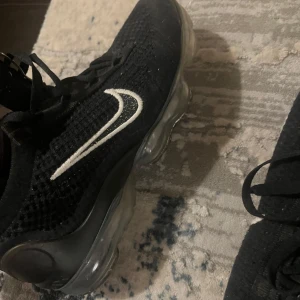 Svarta Nike Vapormax sneakers - Säljer ett par svarta Nike Vapormax sneakers med vit swoosh och transparent sula. Skorna har en stickad ovandel och snörning för en bekväm passform. Perfekta för en sportig look. Använda 2-3 gånger. Köpte dem för 1800kr på rea. 