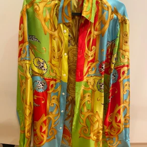Färgglad skjorta från Versace - Säljer en färgglad och iögonfallande skjorta från Versace med ett detaljerat barockmönster i guld, rött, grönt och blått. Skjortan har långa ärmar och knappar framtill. Perfekt för den som vill sticka ut!
