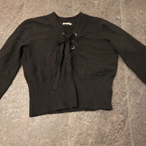 Svart stickad tröja med knytning - Säljer en svart stickad tröja med knytning framtill. Tröjan har långa ärmar och ribbade detaljer vid ärmslut och nederkant. Perfekt för kyligare dagar.