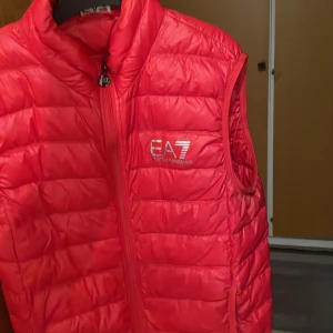 Röd dunväst från Emporio Armani - Snygg röd dunväst från Emporio Armani med dragkedja framtill. Perfekt för kyligare dagar och ger en sportig look. Västen har en quiltad design och är lätt att matcha med olika outfits.