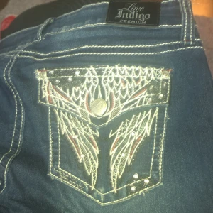 Blå jeans med broderade fickor från Love Indigo - Snygga blå jeans från Love Indigo med unika broderade fickor. Liknar Miss Me jeans. Generellt i väldigt bra skick, tyvärr lite slitna längst ner men enkelt fixat med en ny söm. Storlek 10 motsvarar ungefär storlek 40 i Sverige.