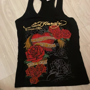 Svart linnetopp från Ed Hardy - Snygg svart linnetopp från Ed Hardy med ett stort, färgglatt tryck av rosor och texten 'Dedicated to the'. Toppen har ett klassiskt Ed Hardy-design med detaljer i rött och guld. Perfekt för en unik och stilfull look.