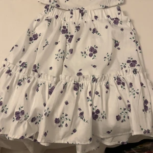 Vit blommig kjol - Charmig vit kjol med lila blommönster och volangdetaljer. Perfekt för en somrig look med sin luftiga design och elastiska midja. Och en matchande topp till.