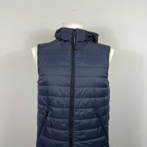 Blå dunväst från C.P. Company - Snygg blå dunväst från C.P. Company med huva och unika glasögondetaljer. Västen har dragkedja framtill och två praktiska fickor med dragkedjor. Perfekt för kyligare dagar. Köpt på Zgander i Helsingborg.