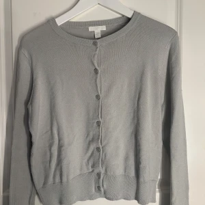 Grå kofta från H&M - Säljer en stilren grå kofta från H&M med knappar framtill. Koftan har långa ärmar och en rund halsringning, perfekt för lager på lager-stil. Passar till både vardag och mer uppklädda tillfällen.