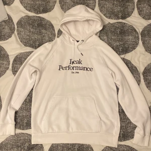 Vit hoodie från Peak Performance - Säljer en stilren vit hoodie från Peak Performance med svart text på framsidan. Väldigt fräsch och bra för en avslappnande look. Den är använd ungefär 3 gånger, inga skador, fläckar eller defekter, helt felfri. Är i storlek XL men passar även L. 