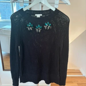 Svart stickad tröja med dekorativa stenar - Säljer en svart stickad tröja med unika dekorativa stenar i turkos och silver på framsidan. Tröjan har långa ärmar och en ribbad kant vid halsen. Perfekt för att ge en touch av glamour till din garderob.
