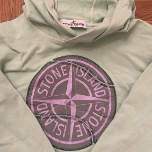 Grön hoodie från Stone Island Junior storlek 10-11år - Snygg ljusgrön hoodie från Stone Island Junior med stort logotryck i lila på framsidan. Perfekt för en avslappnad stil. Mjuk och bekväm med långa ärmar och huva.