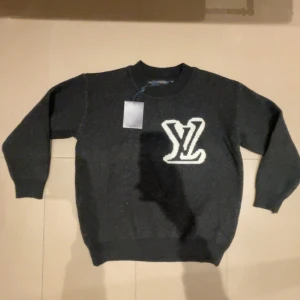 Svart tröja från Louis Vuitton - Snygg svart tröja från Louis Vuitton med ett stort vitt LV-märke på framsidan och baksidan . Tröjan har en klassisk rund hals och långa ärmar, perfekt för en stilren look.