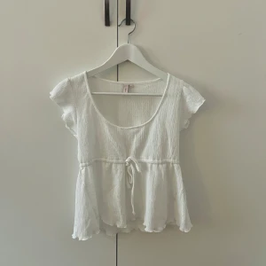 Vit ribbad topp med knyt - Säljer en söt vit ribbad topp med korta ärmar och knytband i midjan. Toppen har en rund halsringning och volangdetaljer vid nederkanten. Perfekt för en somrig look!