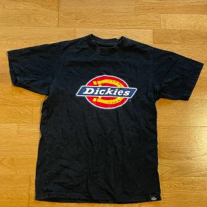 Svart t-shirt från Dickies - Säljer en svart t-shirt från Dickies med det klassiska logotyptrycket i rött, gult och vitt på framsidan. T-shirten är kortärmad och i normal passform, perfekt för en avslappnad stil.