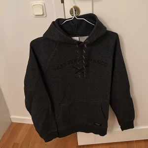 Mörkgrå hoodie från Peak Performance - Säljer en mörkgrå hoodie från Peak Performance med justerbar huva och känguruficka. Perfekt för kyliga dagar och avslappnade stunder. Den har en diskret logga på bröstet och är tillverkad i mjukt material.