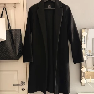 Svart kappa från Zara - Elegant svart kappa från Zara med klassisk design. Långärmad med en stilren siluett som passar till många olika outfits. Vill tillägga att det är ifyllt med rött på zaralappen för att jag skulle veta att det var min, annars inga andra defekter. Storlek S