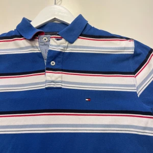 Blå och vit randig pikétröja från Tommy Hilfiger - Snygg blå och vit randig pikétröja från Tommy Hilfiger med röda detaljer. Tröjan har en klassisk krage och korta ärmar, perfekt för en avslappnad stil. Den ikoniska loggan syns på bröstet.