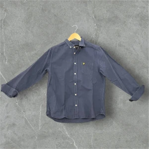 Mörkblå skjorta med knappdetaljer - Snygg mörkblå skjorta med klassisk krage och knappar framtill. Skjortan har en bröstficka med en gul broderad logotyp. Perfekt för en stilren look.