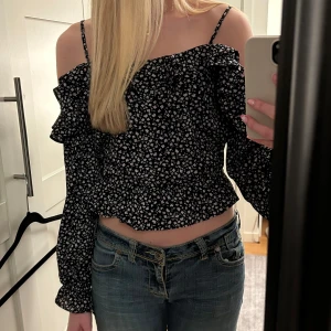 Svart blommig offshoulder blus - Snygg svart offshoulder blus med små vita och rosa blommor. Toppen har långa ärmar och volangdetaljer vid axlarna. Perfekt för en somrig look!