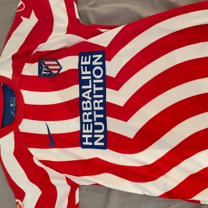 Rödvit randig fotbollströja från Nike - Snygg röd och vit randig fotbollströja från Nike med korta ärmar. Tröjan har ett tryck av 'Herbalife Nutrition' på framsidan och spelarens namn och nummer 'Griezmann 8' på baksidan. Perfekt för fotbollsfans!