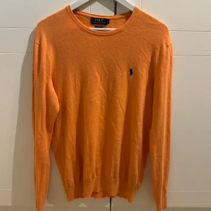 Orange tröja från Polo Ralph Lauren - Snygg orange tröja från Polo Ralph Lauren med det klassiska logotypbroderiet på bröstet. Tröjan har en rund halsringning och långa ärmar, perfekt för en stilren look.