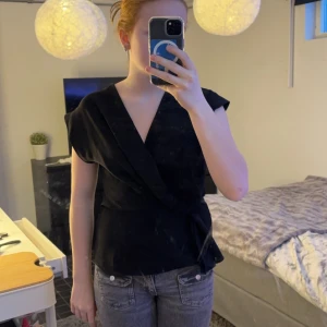 Svart omlottblus från Zara - Snygg svart omlottblus från Zara med knytning i midjan. Blusen har en v-ringad design och korta ärmar, perfekt för en stilren look.