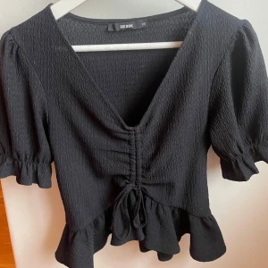 Svart topp från Bik Bok - Snygg svart topp från Bik Bok med v-ringning och volangdetaljer. Toppen har korta ärmar och en knytning framtill som ger en fin passform. Säljer då den är för liten🖤
