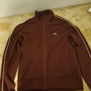 Lacoste tröja - Säljer en speciell röd färgad tröja från Lacoste med dragkedja och vita detaljer längs ärmarna. Tröjan har en hög krage och är i regular fit. Perfekt för en stilren look.