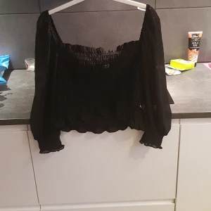 Svart offshoulder topp från Divided - Snygg svart offshoulder topp från Divided med smockdetaljer och långa ärmar. Perfekt för en trendig look med volangdetaljer vid ärmslut och nederkant. Passar bra till både jeans och kjol.