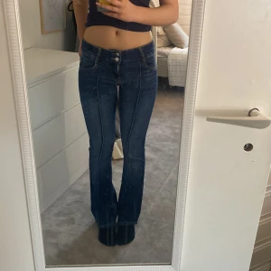 Blå bootcut jeans från Bershka - Snygga blå bootcut jeans från Bershka med dubbla knappar och dragkedja. De har en klassisk femficksdesign och är perfekta för en avslappnad stil. De är lågmidjade och har en söm i mitten.