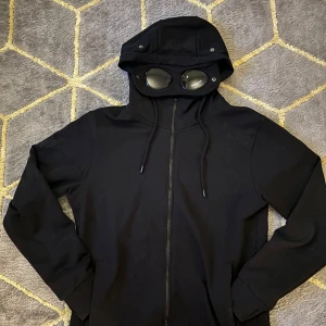 Svart hoodie med glasögon i huvan - Cool svart hoodie med inbyggda glasögon i huvan. Perfekt för en unik stil. Den har dragkedja framtill och långa ärmar. Passar bra för höst och vår.