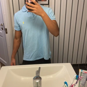 Ljusblå pikétröja från Ralph Lauren - Snygg ljusblå pikétröja från Ralph Lauren med klassisk krage och korta ärmar. Den har en broderad gul logotyp på bröstet. 