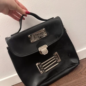 Svart portfölj med metallspänne - Snygg svart portfölj med ett stilrent metallspänne och handtag. Väskan har en praktisk dragkedja inuti för extra säkerhet. Perfekt för att bära dokument eller en liten laptop. 