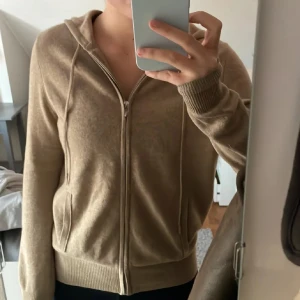 Beige hoodie med dragkedja - Säljer en mysig beige hoodie med dragkedja framtill. Tröjan har långa ärmar och ribbade muddar vid ärmslut och nederkant. Perfekt för en avslappnad stil.