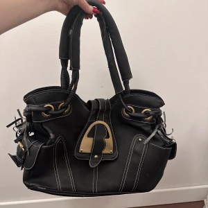 Svart handväska med detaljer - Snygg svart handväska med unika detaljer och kontrasterande sömmar. Väskan har två handtag och flera fack inuti för praktisk förvaring. Perfekt för att bära med sig det nödvändigaste med stil.