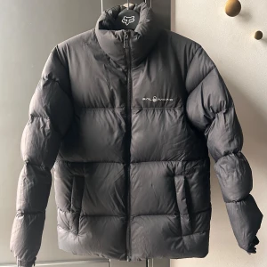 Svart dunjacka från Sail Racing stl 146-152  - Snygg svart dunjacka från Sail Racing med dragkedja och hög krage. Jackan har en pufferdesign och logotyp på bröstet och ryggen. Storlek 146-152.