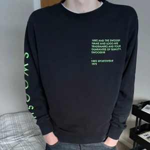 Svart långärmad tröja från Nike - Säljer en svart långärmad tröja från Nike med grön text på bröstet och ärmen. Tröjan har en rund halsringning och är perfekt för en sportig look. Texten 'SWOOSH' finns längs ärmen i en iögonfallande grön färg.