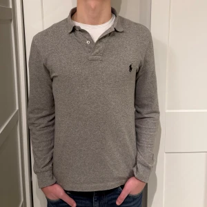 Ralph Lauren pikétröja - Långärmad Ralph Lauren pike i färgen grå. Inga defekter på tröjan. Size S (slim fit) modellen är 177cm. Hör av dig vid funderingar!