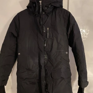 Svart dunjacka från Peak Performance - Snygg och varm svart Vinter dunjacka från Peak Performance med huva och dragkedja. Jackan har långa ärmar och praktiska fickor framtill. Perfekt för kalla dagar.