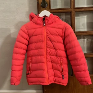 Korallfärgad vinterjacka  från Tuxer - Säljer en snygg korallfärgad vinterjacka från Tuxer med huva och dragkedja. Jackan är perfekt för kyliga dagar och skidåkning. Luvan är avtagbar, den är även lila i luvan och har lila foder, 
