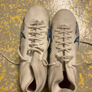 Vita Nike fotbollsskor med blå detaljer - Snygga vita Nike fotbollsskor med blå och rosa detaljer. Skorna har snörning och en modern design med Nike Zoom-teknologi för optimal prestanda på planen.pris kan diskuteras 